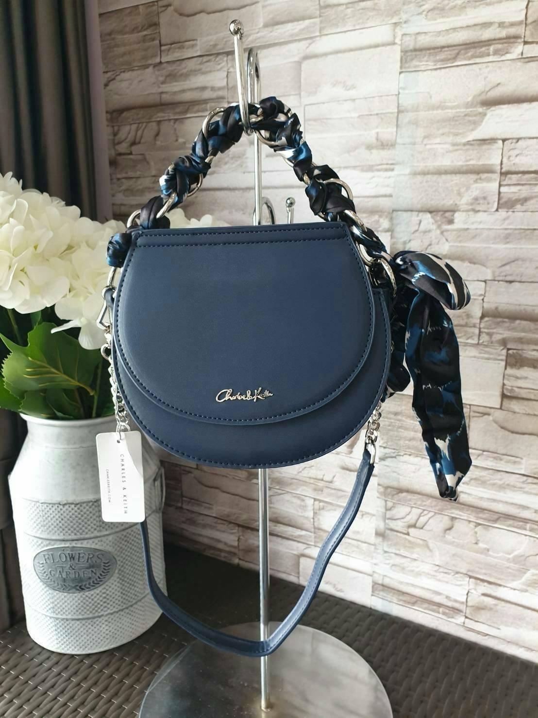 CHARLES & KEITH รุ่น LEATHER SATIN SCARF CROSSBODY BAG SL2-50150828 ราคาปกติช้อป ( ฿3,590.00 ) กระเป๋าถือ และ สะพาย ทรงโค้งมน หนังเรียบ ตั้งอยู่ทรง ด้านหน้าติดโลโก้แบรนด์ อะไหล่เงิน หูจับสายกระเป๋าร้อยเข้ากับห่วงเหล็กกลมๆ ด้วยผ้าลายสวยงามผูกเป็นโบว์เก๋ๆ ถ