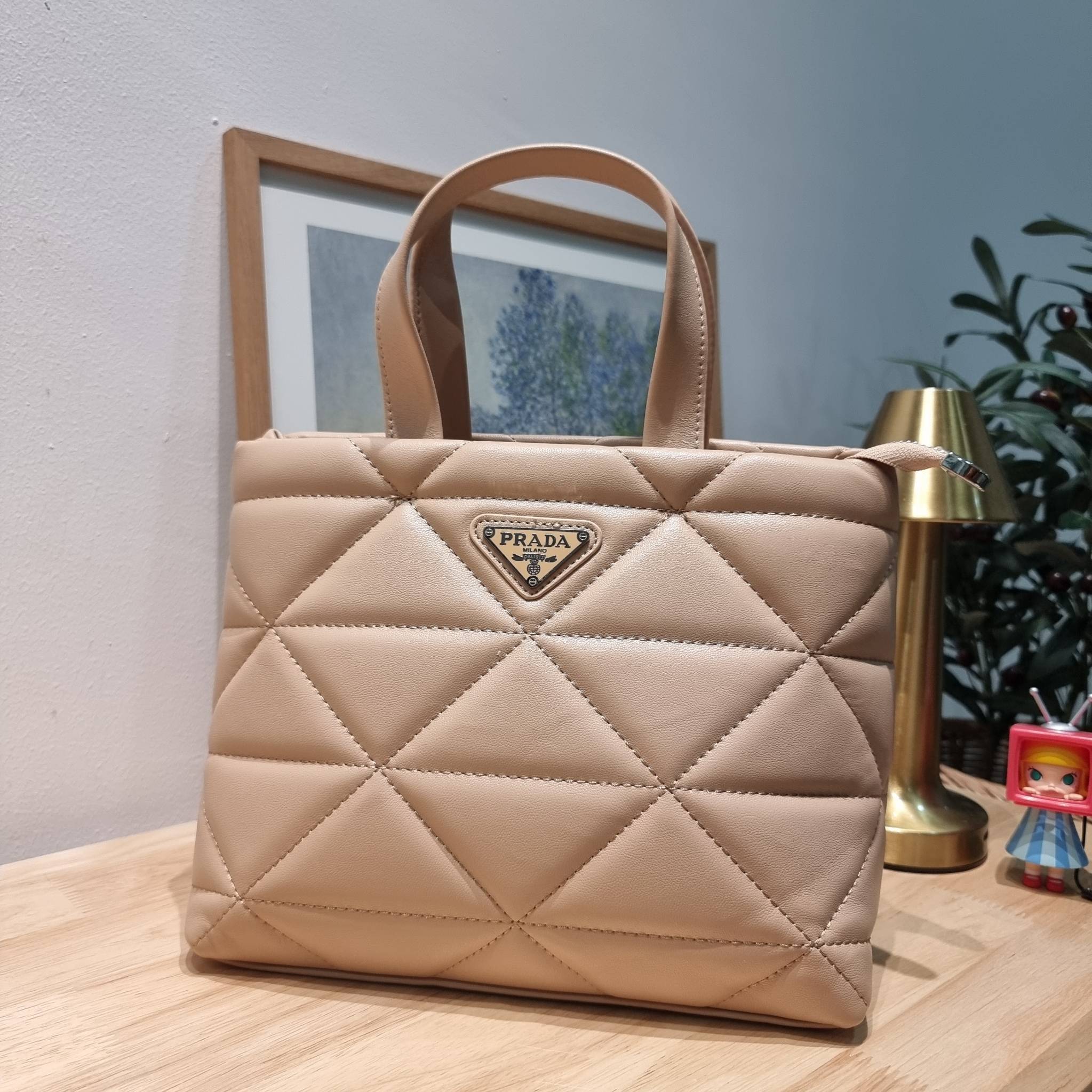 PRADA LEATHER PADDED BAG ใหม่ล่าสุด กับคอลเลคชั่นที่ทำออกมาตอบโจทย์ได้ทุกไลฟ์สไตล์ และเป็นยูนิเซ็กส์ กับกระเป๋าทรงโท้ท ขนาดกำลังดี วัสดุหนังเรียบ ตัดเย็บเดินเส้นลายตาราง ปากกระเป๋ามีซิปเปิดปิด ภายในเป็นช่องโล่ง