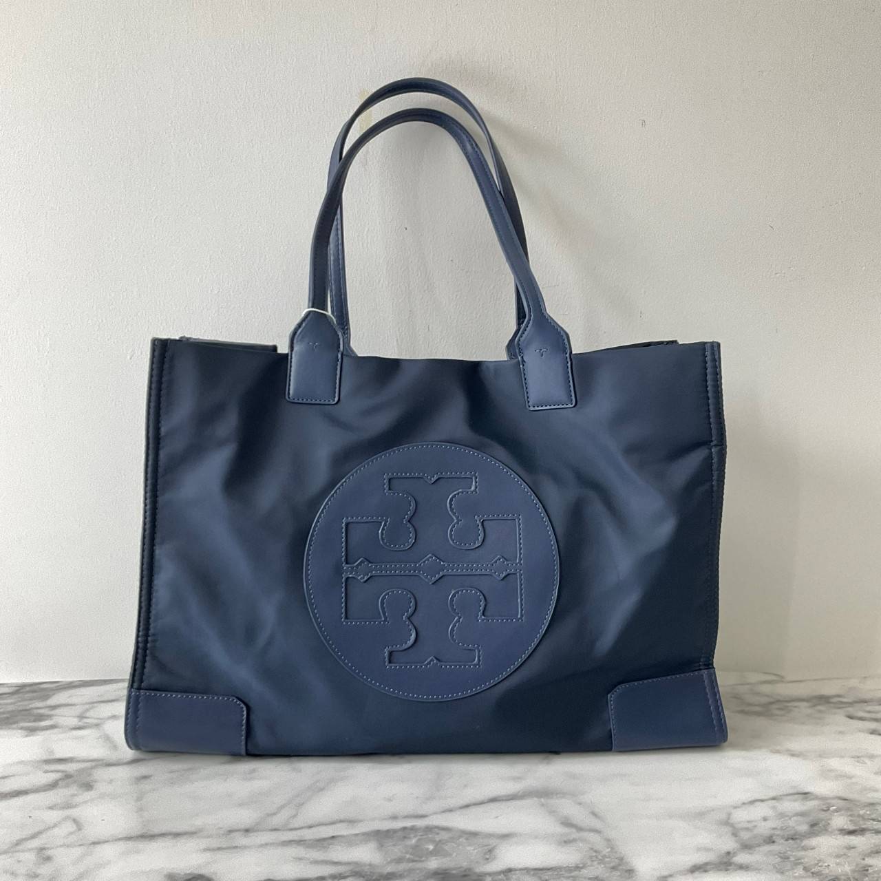 กระเป๋าทรงโท้ทใบใหญ่ TORY BURCH ELLA TOTE BAG วัสดุ NYLON ทั้งใบตัดด้วยหนังสีดำ แต่งโลโก้แบรนด์ด้านหน้า ด้านข้างมีกระดุม สามารถขยายทรงได้ ด้านในเปิดปิดด้วยกระดุมแม่เหล็กฝัง ช่องกว้างหลักหนึ่งช่องและช่องซิปอีกช่อง สามารถใส่แล็ปท็อปได้สบาย ใบใหญ่เว่อวัง ไอเ