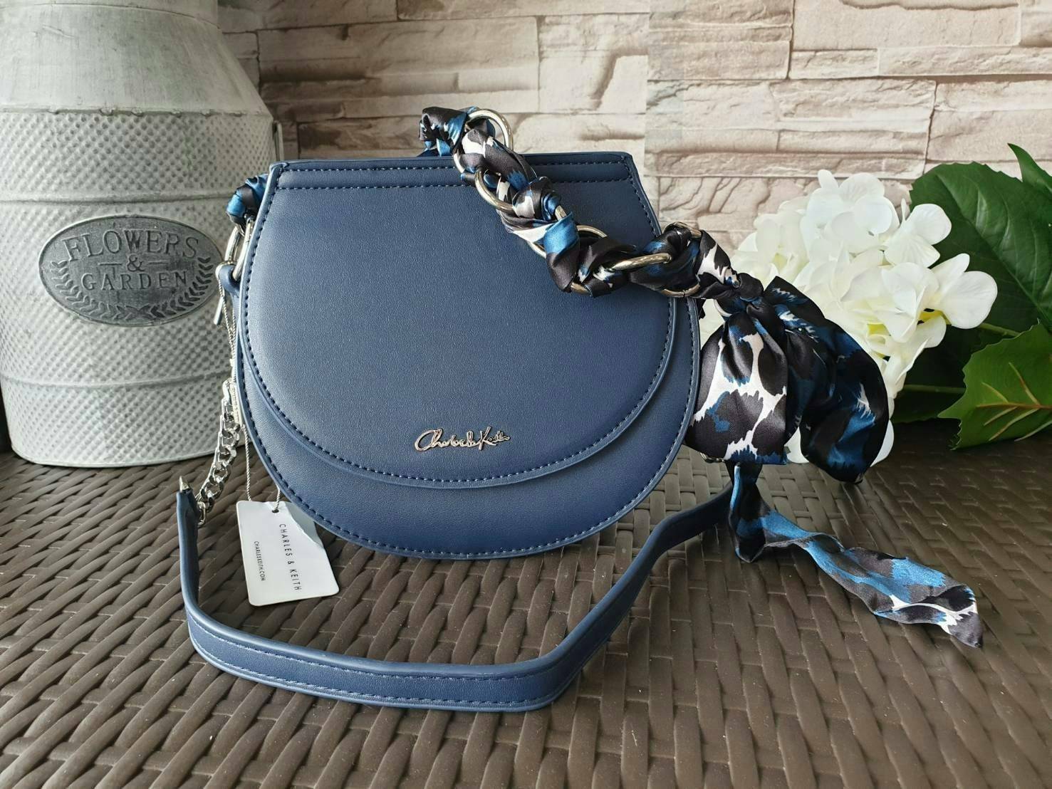 CHARLES & KEITH รุ่น LEATHER SATIN SCARF CROSSBODY BAG SL2-50150828 ราคาปกติช้อป ( ฿3,590.00 ) กระเป๋าถือ และ สะพาย ทรงโค้งมน หนังเรียบ ตั้งอยู่ทรง ด้านหน้าติดโลโก้แบรนด์ อะไหล่เงิน หูจับสายกระเป๋าร้อยเข้ากับห่วงเหล็กกลมๆ ด้วยผ้าลายสวยงามผูกเป็นโบว์เก๋ๆ ถ