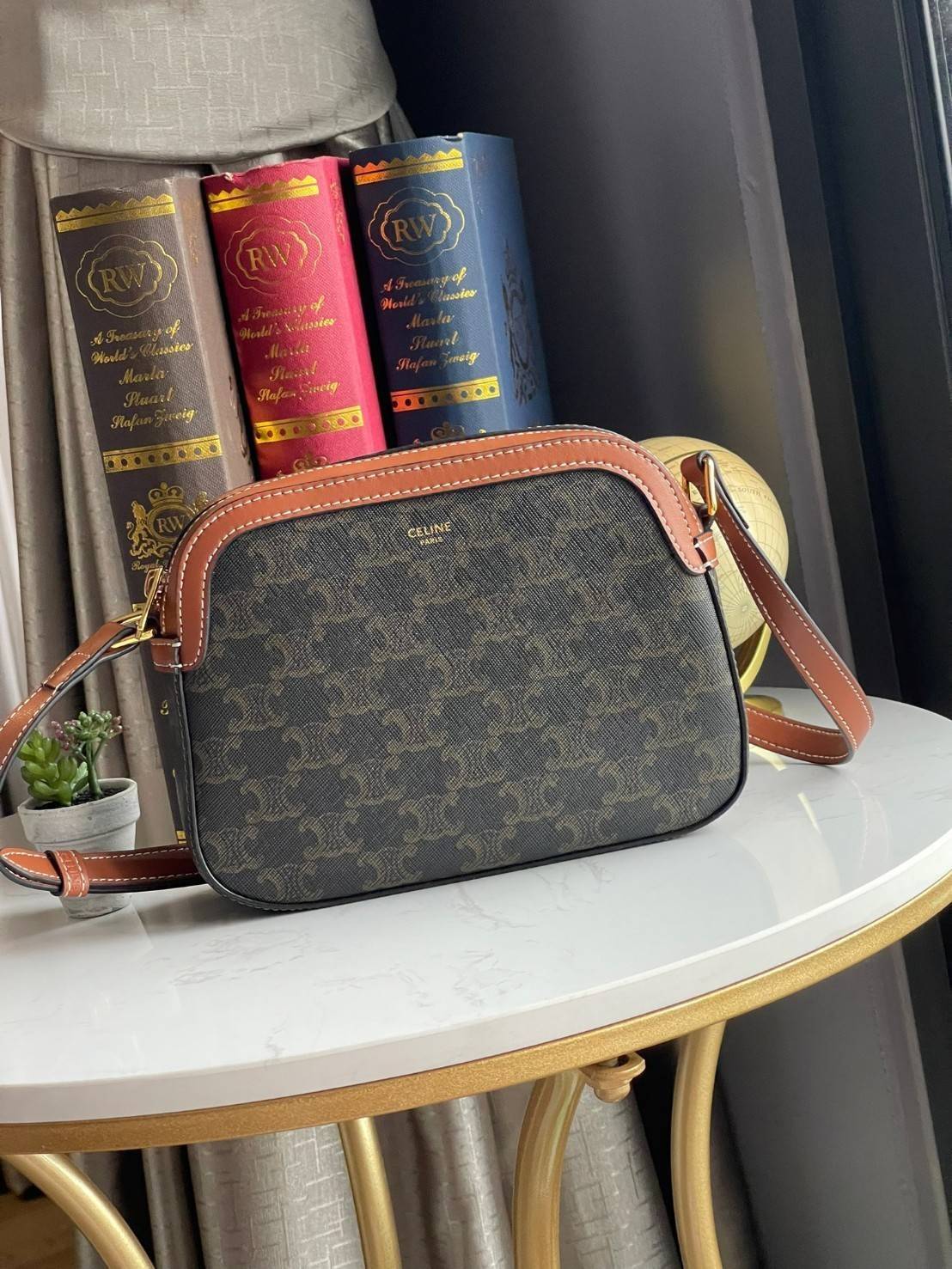 CELINE Triomphe Canvas Small Camera กระเป๋าสะพายไหล่สุดเก๋ใบนี้ผลิตจากผ้าใบเคลือบอย่างดีพร้อมขอบหนังสีน้ำตาล มาพร้อมสายสะพายหนังปรับระดับได้พร้อมฮาร์ดแวร์สีทองและซิปด้านบน ภายในบุด้วยผ้าสีน้ำตาล ลวดลาย triomphe อันเป็นเอกลักษณ์ของแบรนด์ และเป็นกระเป๋าสะพา