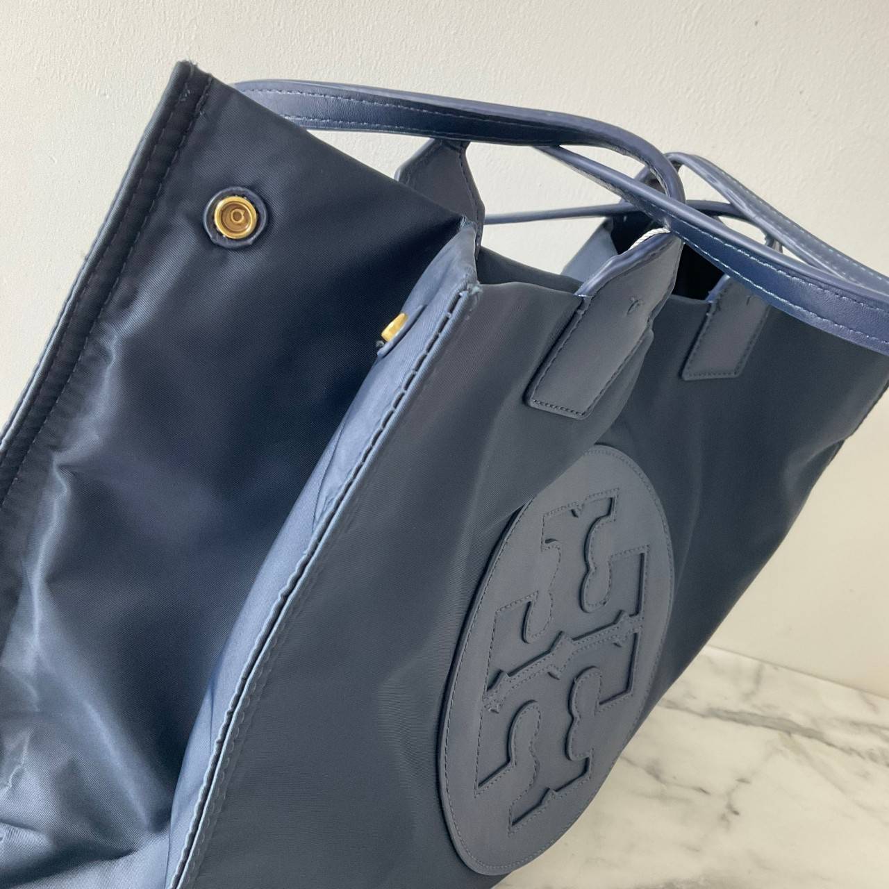 กระเป๋าทรงโท้ทใบใหญ่ TORY BURCH ELLA TOTE BAG วัสดุ NYLON ทั้งใบตัดด้วยหนังสีดำ แต่งโลโก้แบรนด์ด้านหน้า ด้านข้างมีกระดุม สามารถขยายทรงได้ ด้านในเปิดปิดด้วยกระดุมแม่เหล็กฝัง ช่องกว้างหลักหนึ่งช่องและช่องซิปอีกช่อง สามารถใส่แล็ปท็อปได้สบาย ใบใหญ่เว่อวัง ไอเ