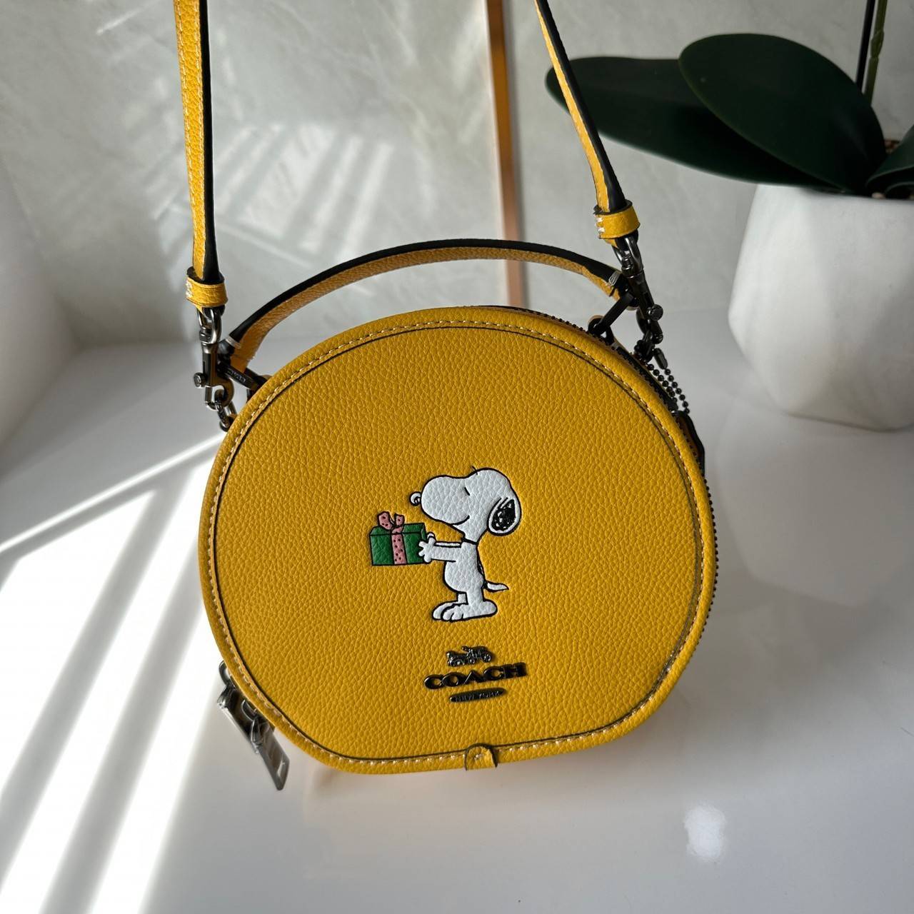 COACH X PEANUTS CANTEEN CROSSBODY WITH SNOOPY PRESENT MOTIF (CE845,CF290) กระเป๋าสะพายข้างทรงกลมไซส์มินิ มีหูจับ วัสหนังแท้ ด้านหน้าโดดเด่นด้วยรูปการ์ตูนSnoopy